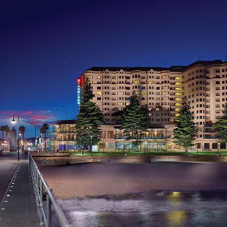 Stamford Grand Adelaide Hotel Glenelg Bagian luar foto The photo shows a waterfront scene at dusk, featuring a large building lit up against a darkening sky. The architecture appears modern and prominent, with several windows illuminated. In the foreground, there is a walkway or pier with fencing along t