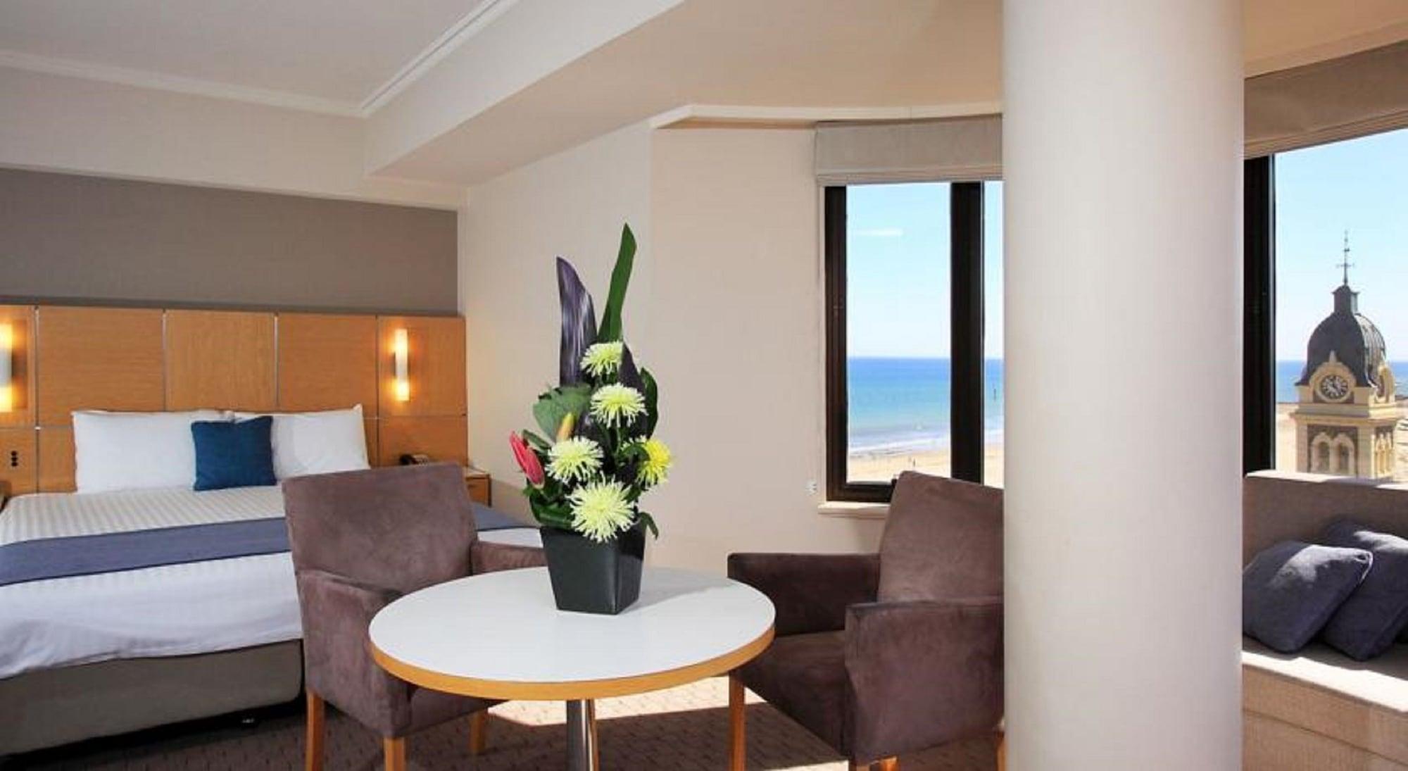 Stamford Grand Adelaide Hotel Glenelg Bagian luar foto The photo shows a cozy hotel room with a comfortable seating arrangement. There are two brown chairs positioned around a round table, which features a decorative flower arrangement in a black vase. The background includes a window that offers a view 