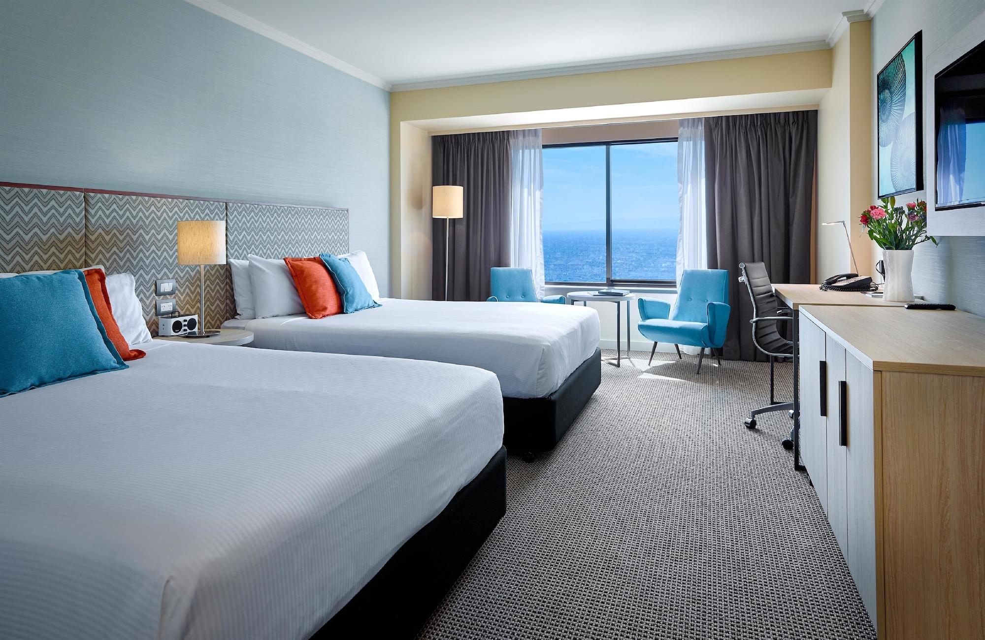 Stamford Grand Adelaide Hotel Glenelg Bagian luar foto The photo shows a well-decorated hotel room featuring two queen-sized beds with white linens and colorful accent pillows. There is a large window with curtains that offers a view of the ocean. A small seating area with a blue chair and a matching des