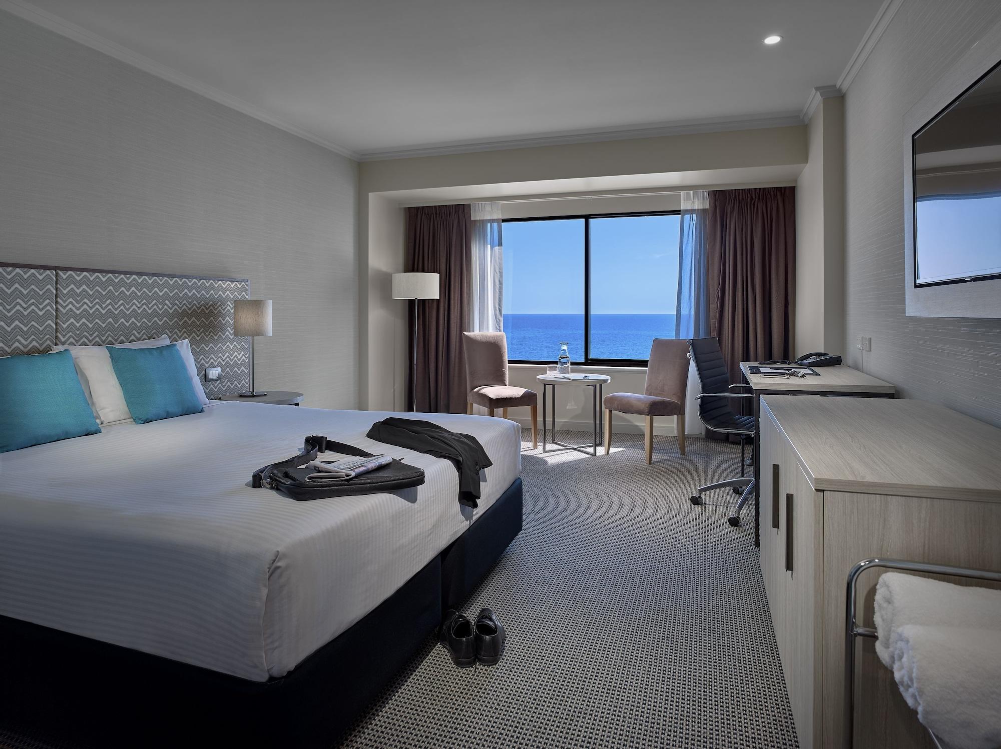 Stamford Grand Adelaide Hotel Glenelg Bagian luar foto The photo shows a modern hotel room featuring a queen-sized bed with white linens and two aqua blue decorative pillows. There are bedside lamps and a large window with a view of the ocean. The room also has a small seating area with two chairs and a 