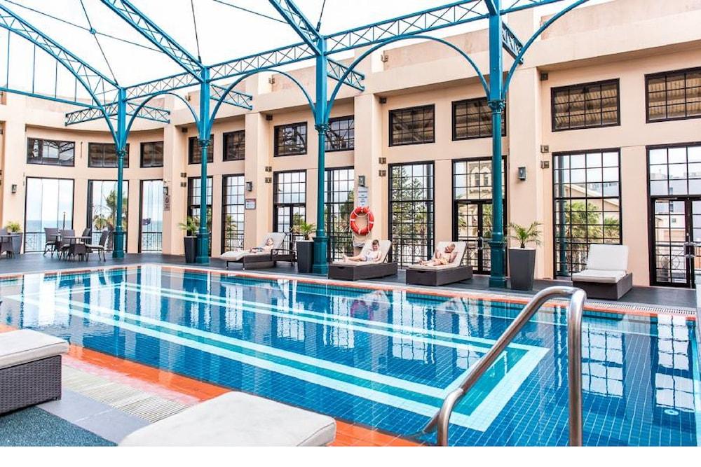 Stamford Grand Adelaide Hotel Glenelg Bagian luar foto The photo shows an indoor swimming pool area characterized by a modern and elegant design. The pool features blue tiles with two white stripes running along its length. Surrounding the pool, there are several lounge chairs for relaxation. The walls h
