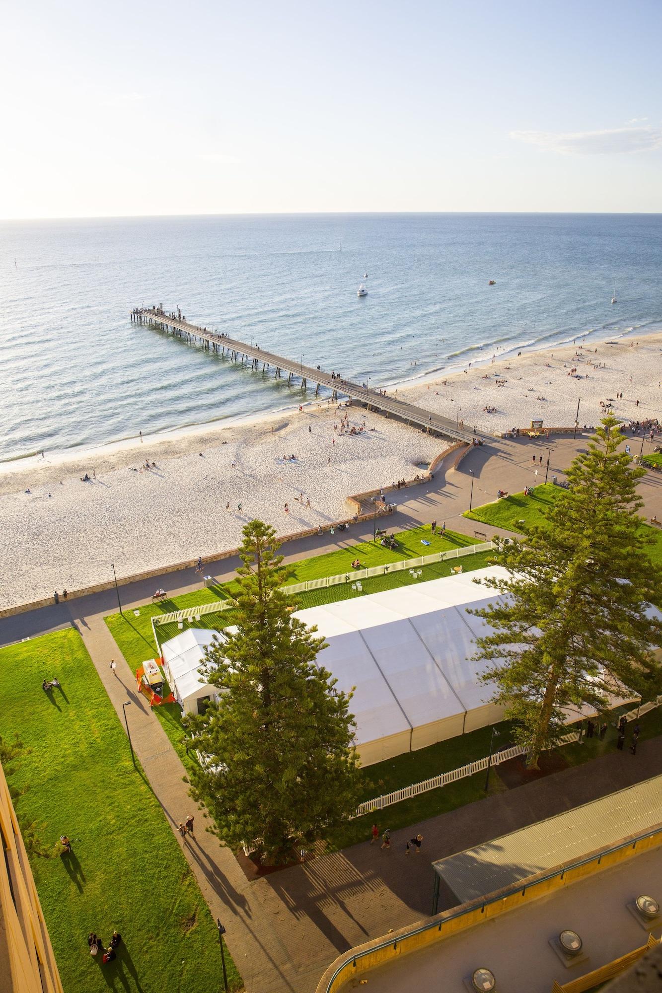 Stamford Grand Adelaide Hotel Glenelg Bagian luar foto The photo shows a scenic view of a beach with a long pier extending into the water. The beach is sandy, and there are people enjoying the sun and walking along the shore. In the foreground, there is a large white tent set up on a grassy area, likely 