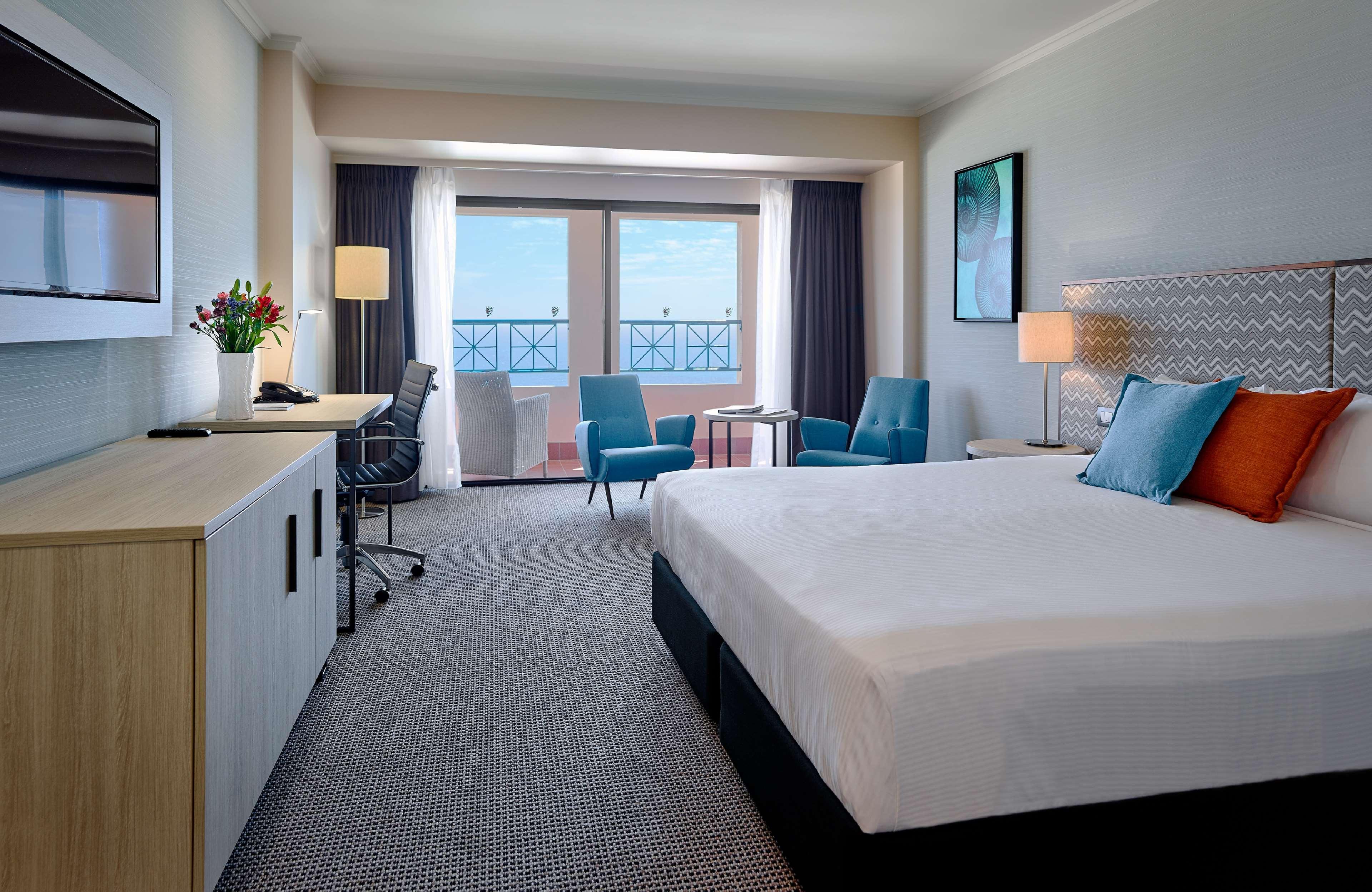 Stamford Grand Adelaide Hotel Glenelg Bagian luar foto The photo depicts a modern hotel room. The room features a large bed with a white comforter, and there are decorative pillows in orange and blue. There is a flat-screen TV mounted on the wall, and a desk with an office chair is positioned near the wi