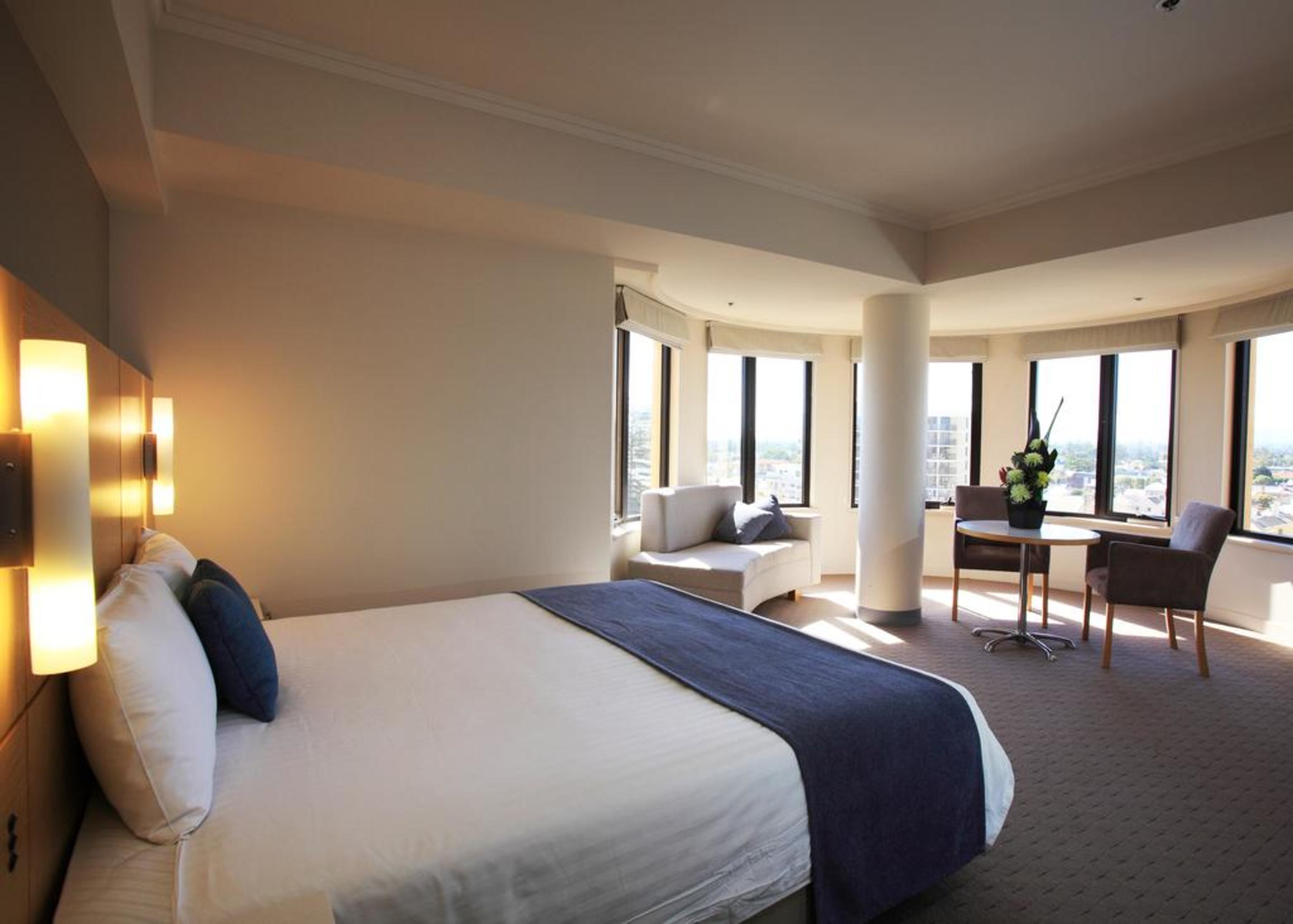 Stamford Grand Adelaide Hotel Glenelg Bagian luar foto The photo shows a hotel room with a modern and spacious design. There is a large bed with white linens and a dark blue throw blanket. The room features multiple windows that allow plenty of natural light, and there is a small seating area near the wi