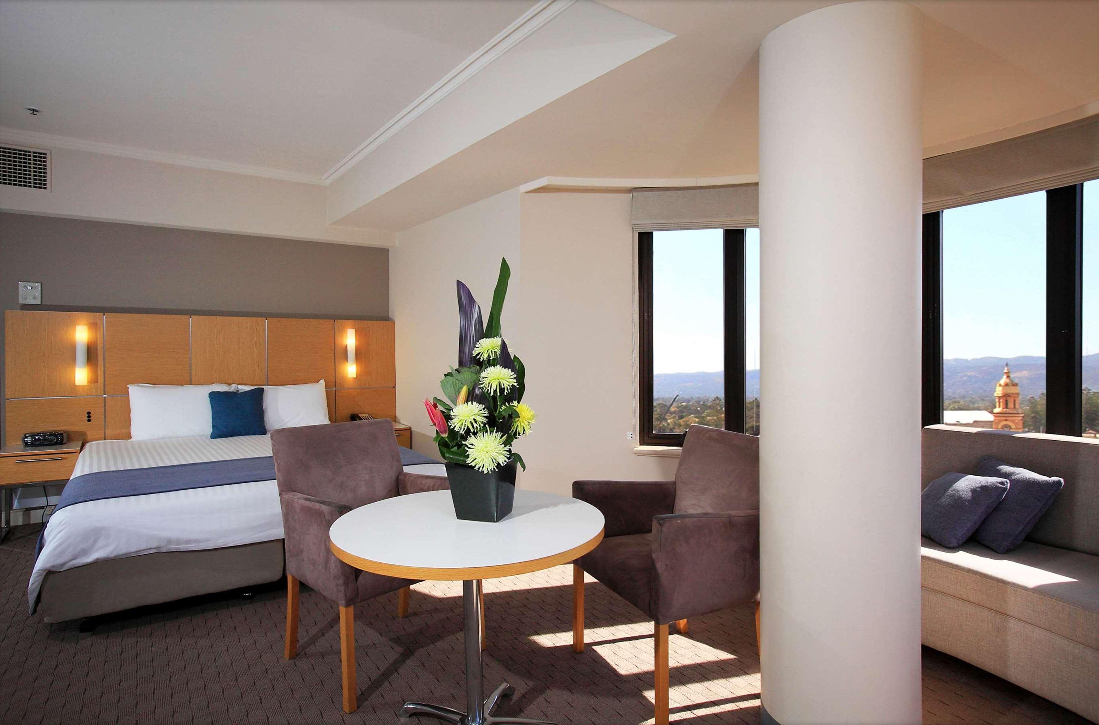 Stamford Grand Adelaide Hotel Glenelg Bagian luar foto The photo shows a hotel room with a modern design. On one side, there is a plush bed with white and blue bedding. In the center of the room, there's a small round table with two gray upholstered chairs around it. A vase of flowers sits on the table, 