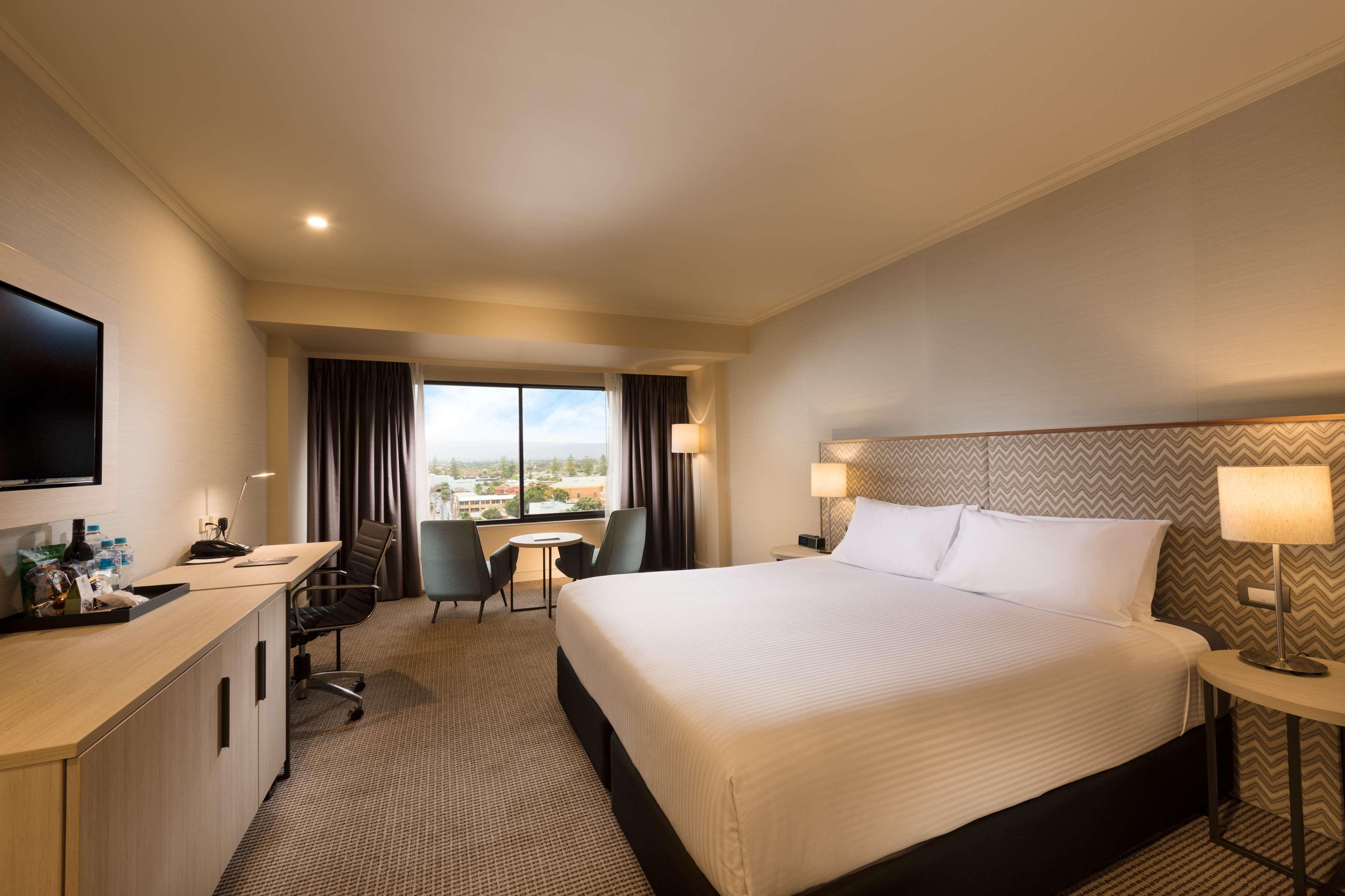Stamford Grand Adelaide Hotel Glenelg Bagian luar foto The photo shows a modern hotel room. It features a large bed with white linens, a desk with a chair, and a flat-screen TV mounted on the wall. There are two armchairs and a small table near a large window that lets in natural light, providing a view 