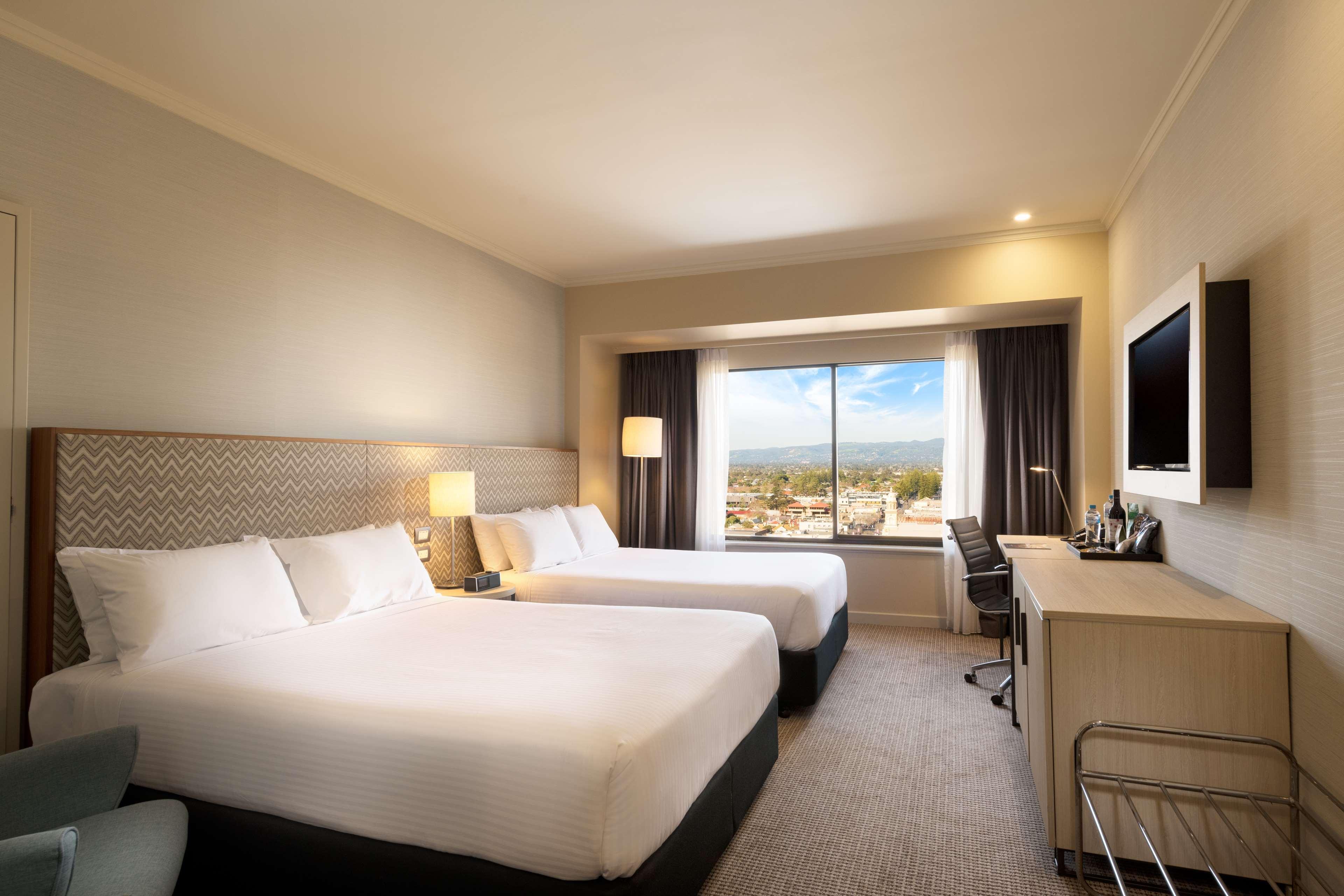 Stamford Grand Adelaide Hotel Glenelg Bagian luar foto The photo shows a modern hotel room featuring two beds with white linens. There are bedside tables with lamps next to each bed. A large window provides natural light and offers a view of the outside, with curtains on either side. The room has a neutr