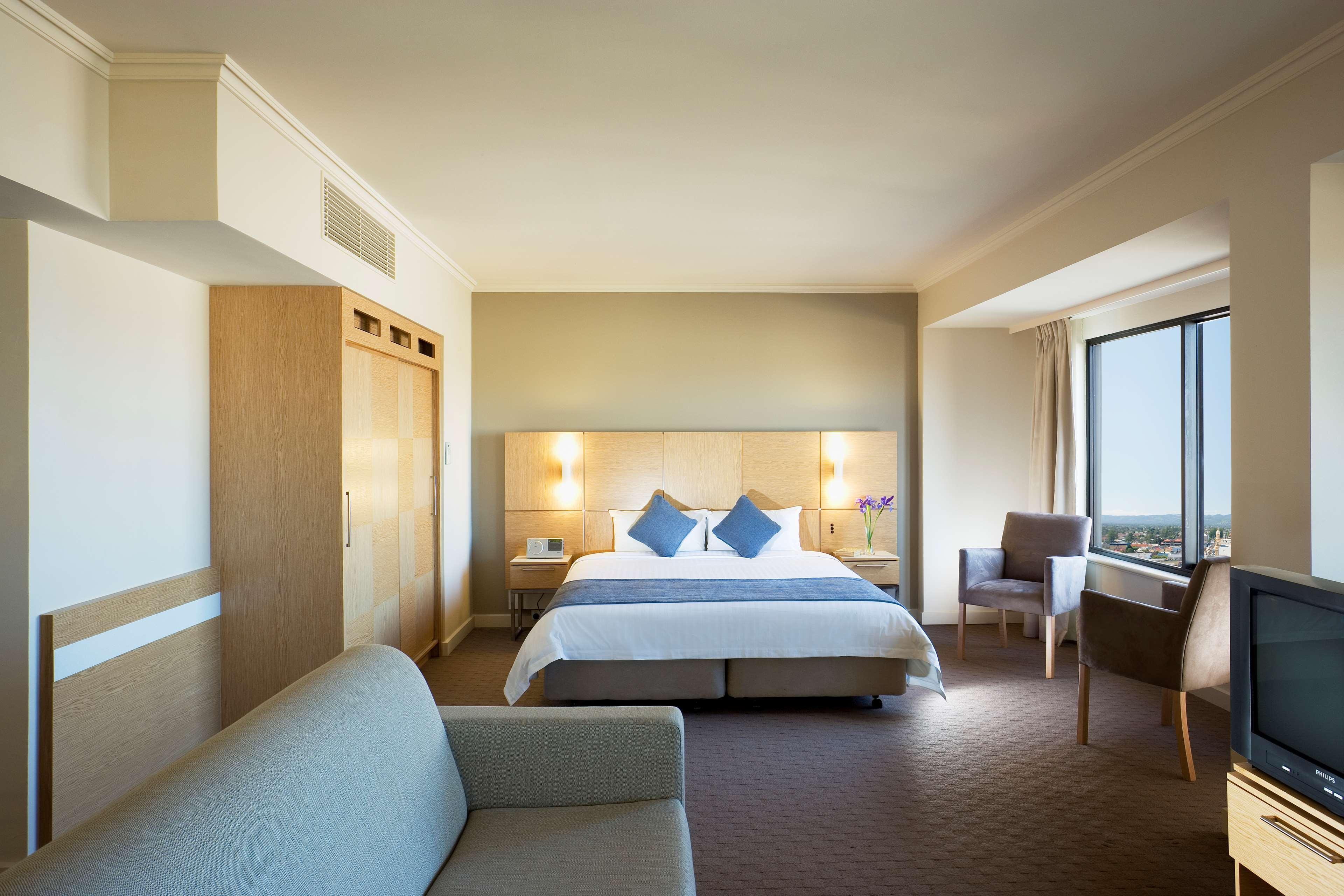Stamford Grand Adelaide Hotel Glenelg Ruang foto The photo shows a modern hotel room featuring a large bed with two blue decorative pillows. There are bedside lamps on both sides of the headboard. To the left, there's a wooden wardrobe and a small table with a vase and a few amenities. On the right