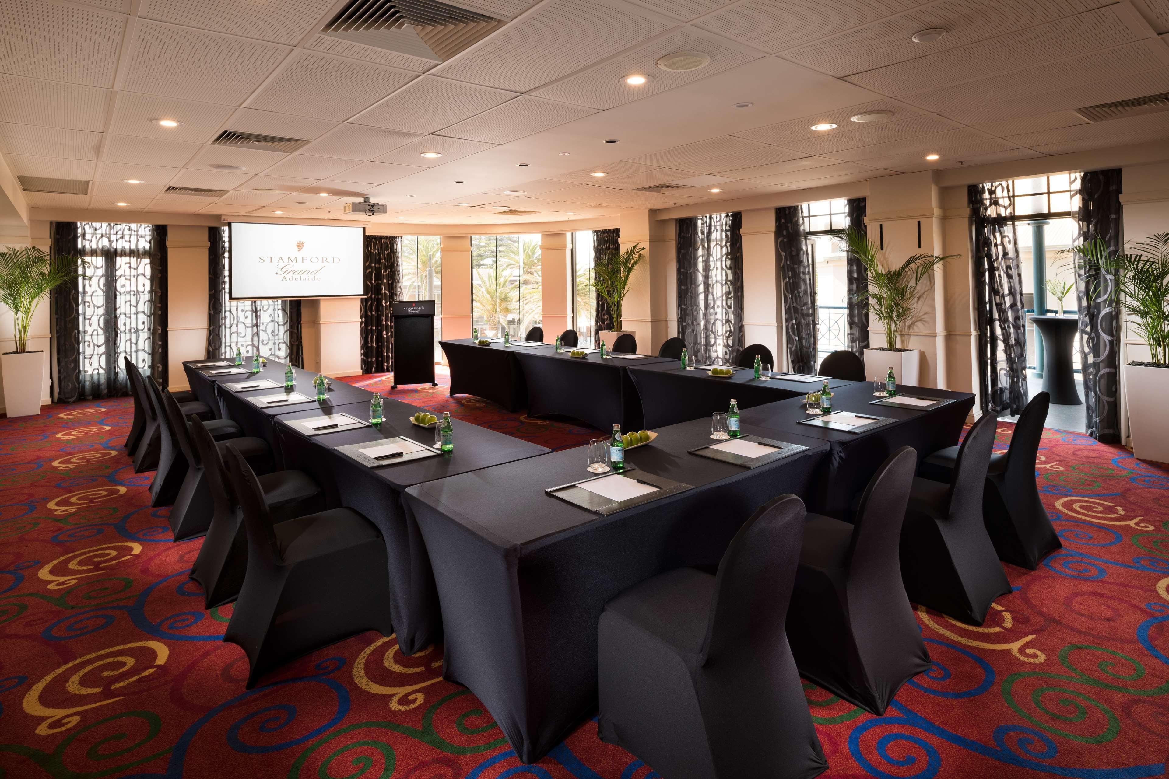 Stamford Grand Adelaide Hotel Glenelg Bagian luar foto The photo shows a conference room set up for a meeting or presentation. There are several tables arranged in a U-shape, covered with black tablecloths. Each table has a notepad and a small bottle of water on it. The chairs around the tables are also 