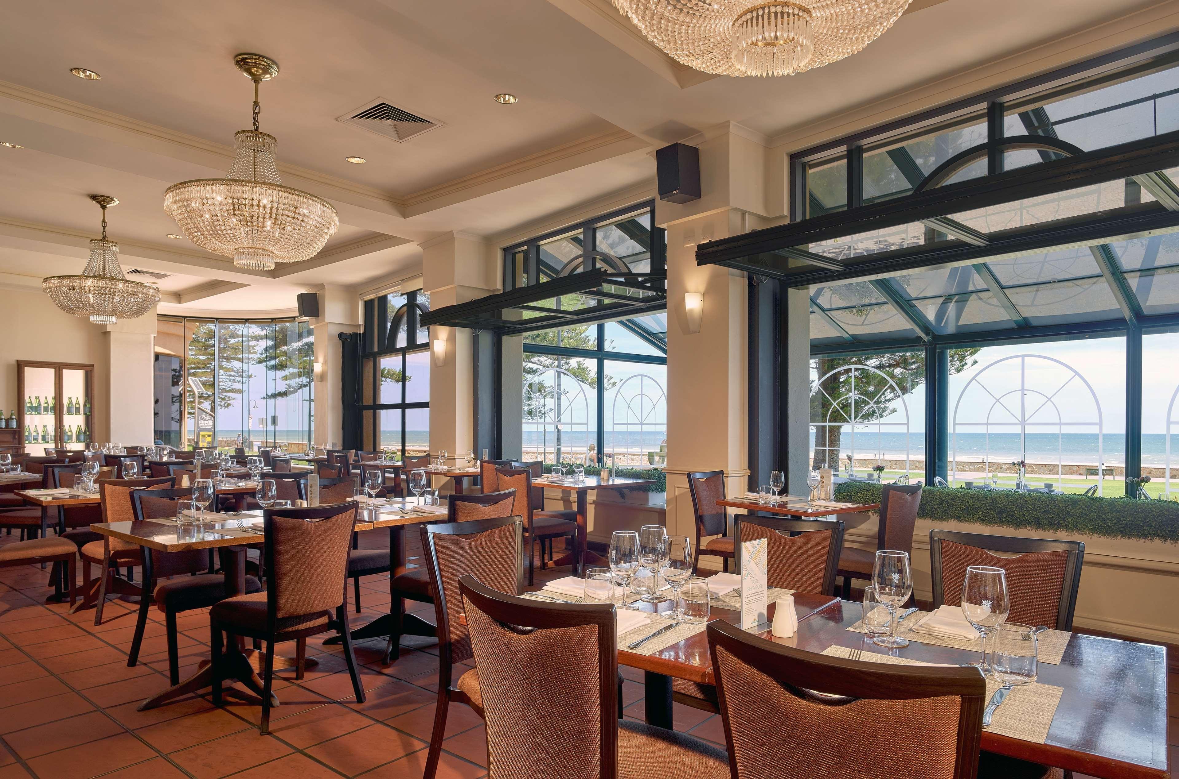 Stamford Grand Adelaide Hotel Glenelg Bagian luar foto The photo shows the interior of a restaurant. It features several wooden tables set with glasses and utensils, suggesting it's ready for diners. The decor includes elegant chandeliers hanging from the ceiling, adding a touch of sophistication. Large 