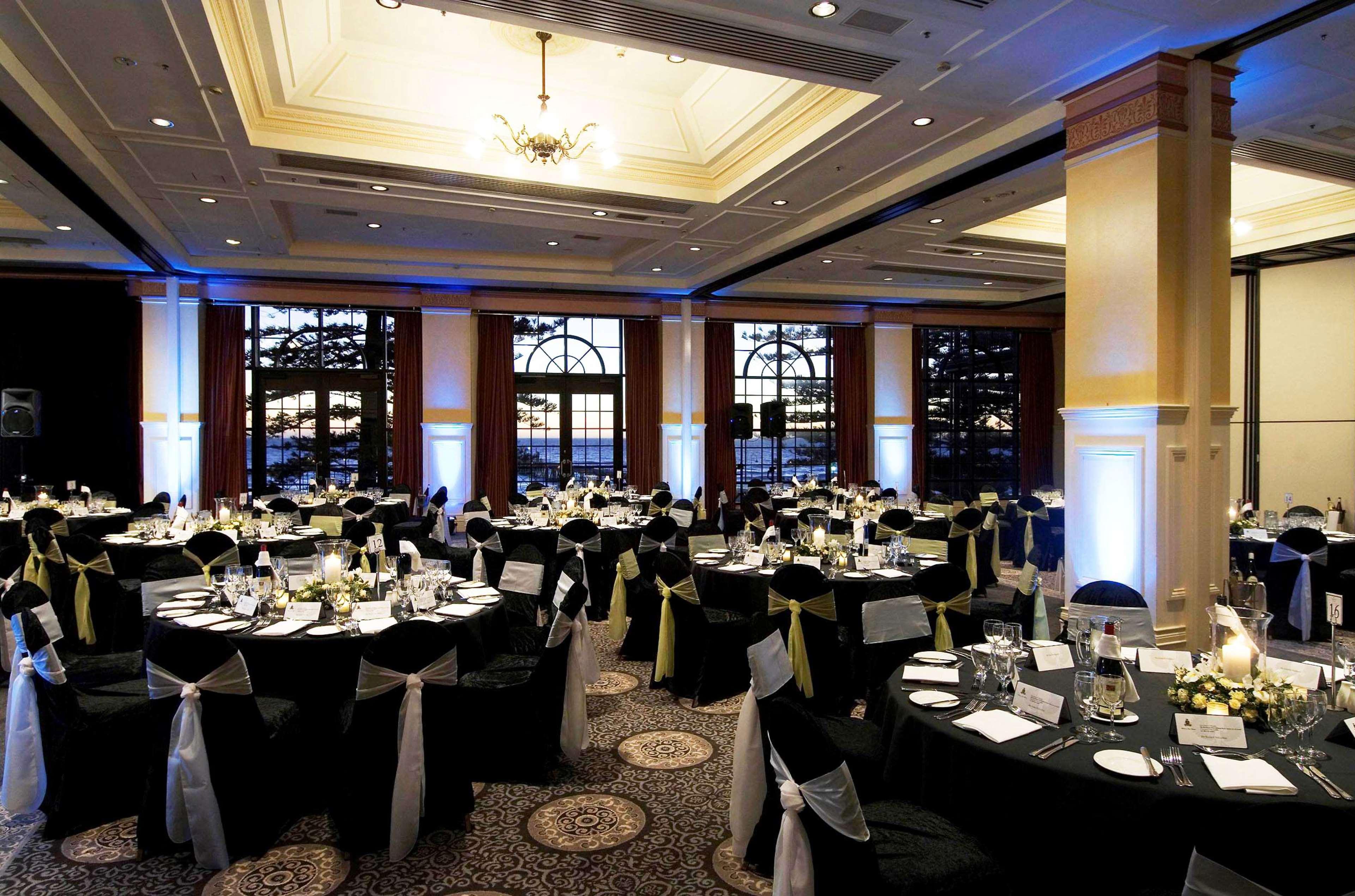 Stamford Grand Adelaide Hotel Glenelg Restoran foto The photo depicts an elegantly arranged banquet hall set for an event. The room features round tables covered with black tablecloths, each adorned with white tableware and floral centerpieces. Chairs are draped in white fabric with decorative sashes 
