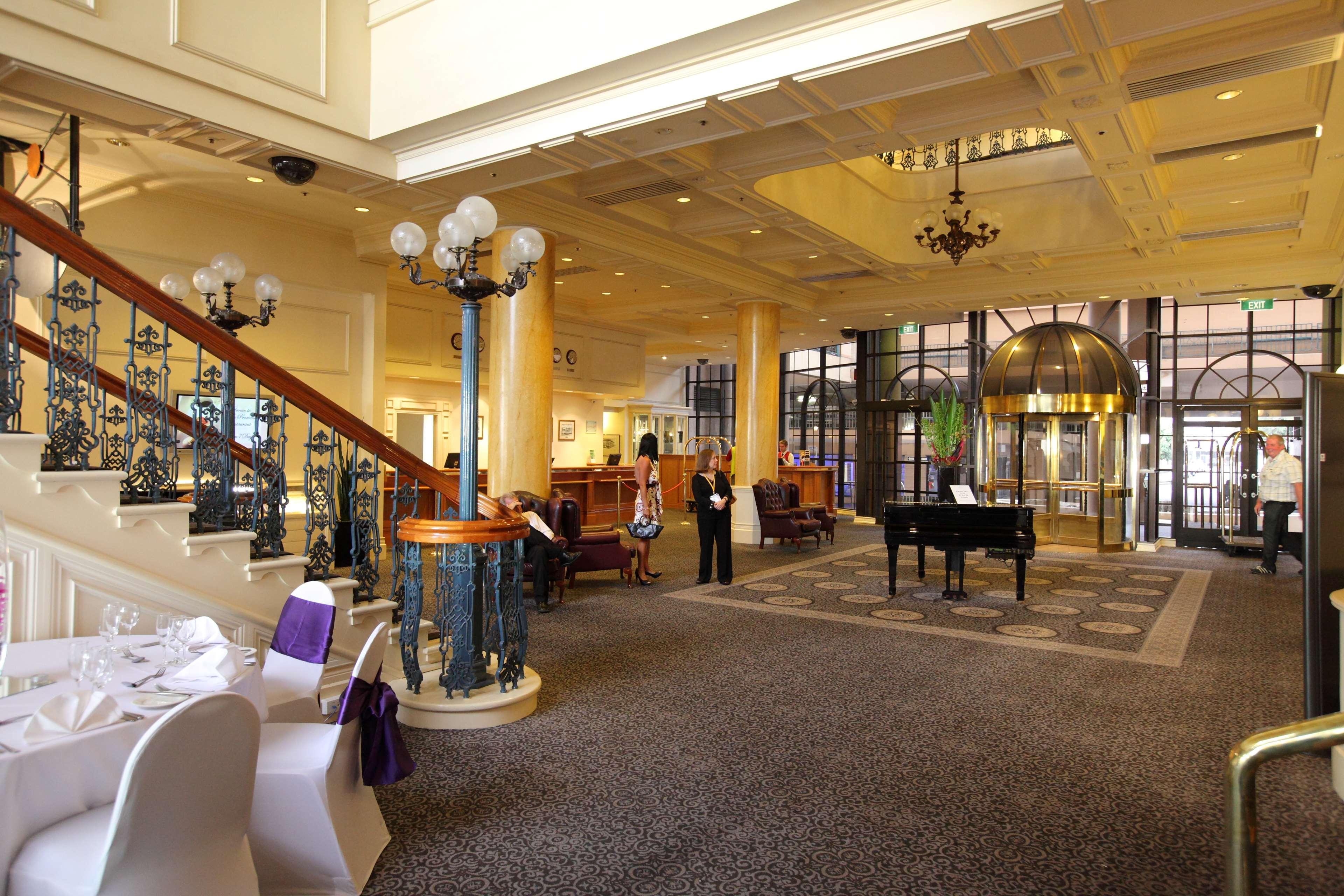 Stamford Grand Adelaide Hotel Glenelg Interior foto The photo shows a luxurious interior of a hotel or a restaurant lobby. There is an elegant staircase with ornate railings leading to an upper level. The space features high ceilings with decorative chandeliers and ample natural light coming through l