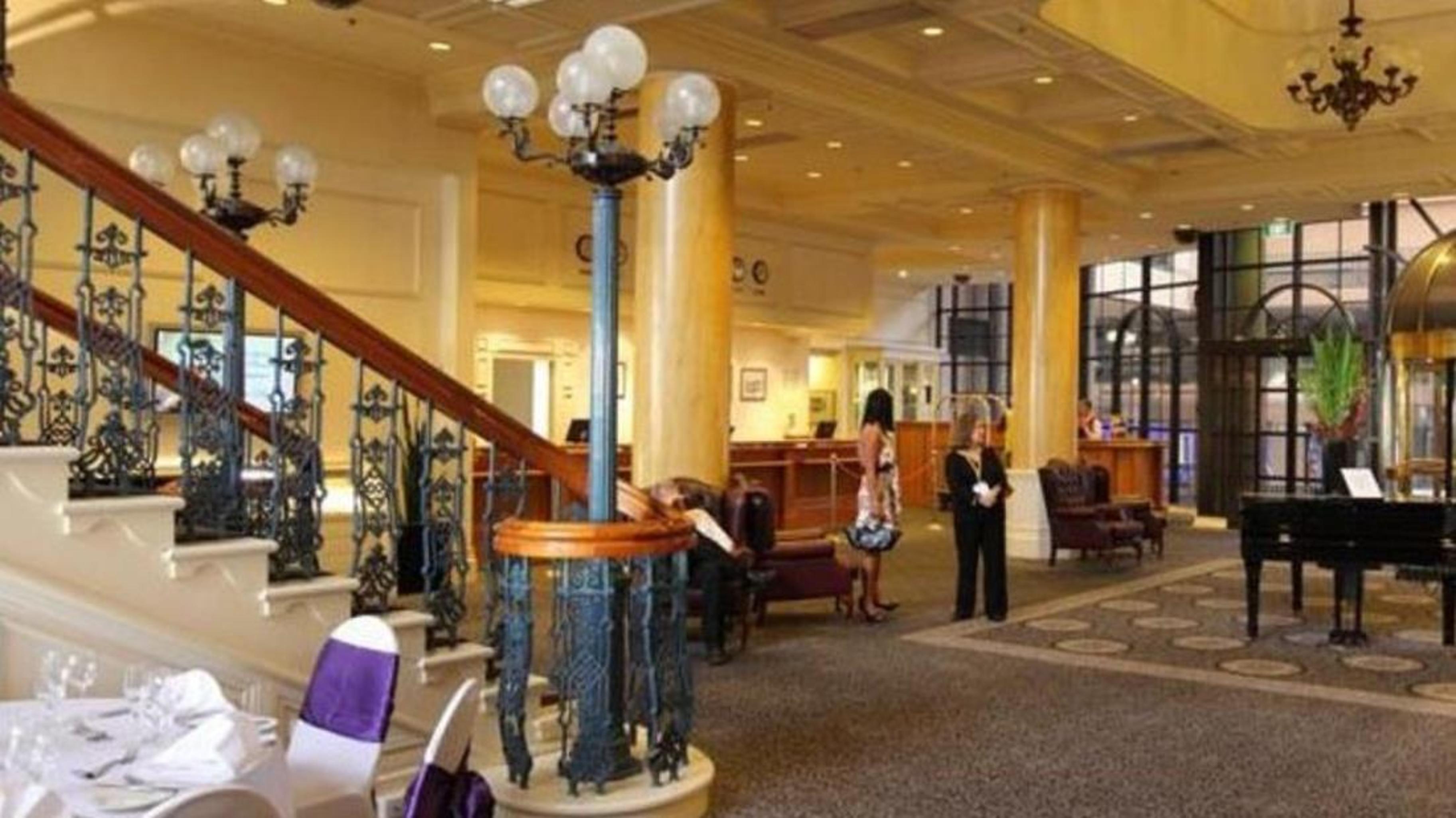 Stamford Grand Adelaide Hotel Glenelg Bagian luar foto The photo shows the interior of a hotel lobby. It features a spacious area with a high ceiling and elegant decor. There is a staircase with a decorative railing leading to an upper level. In the foreground, some guests and staff are present, and ther