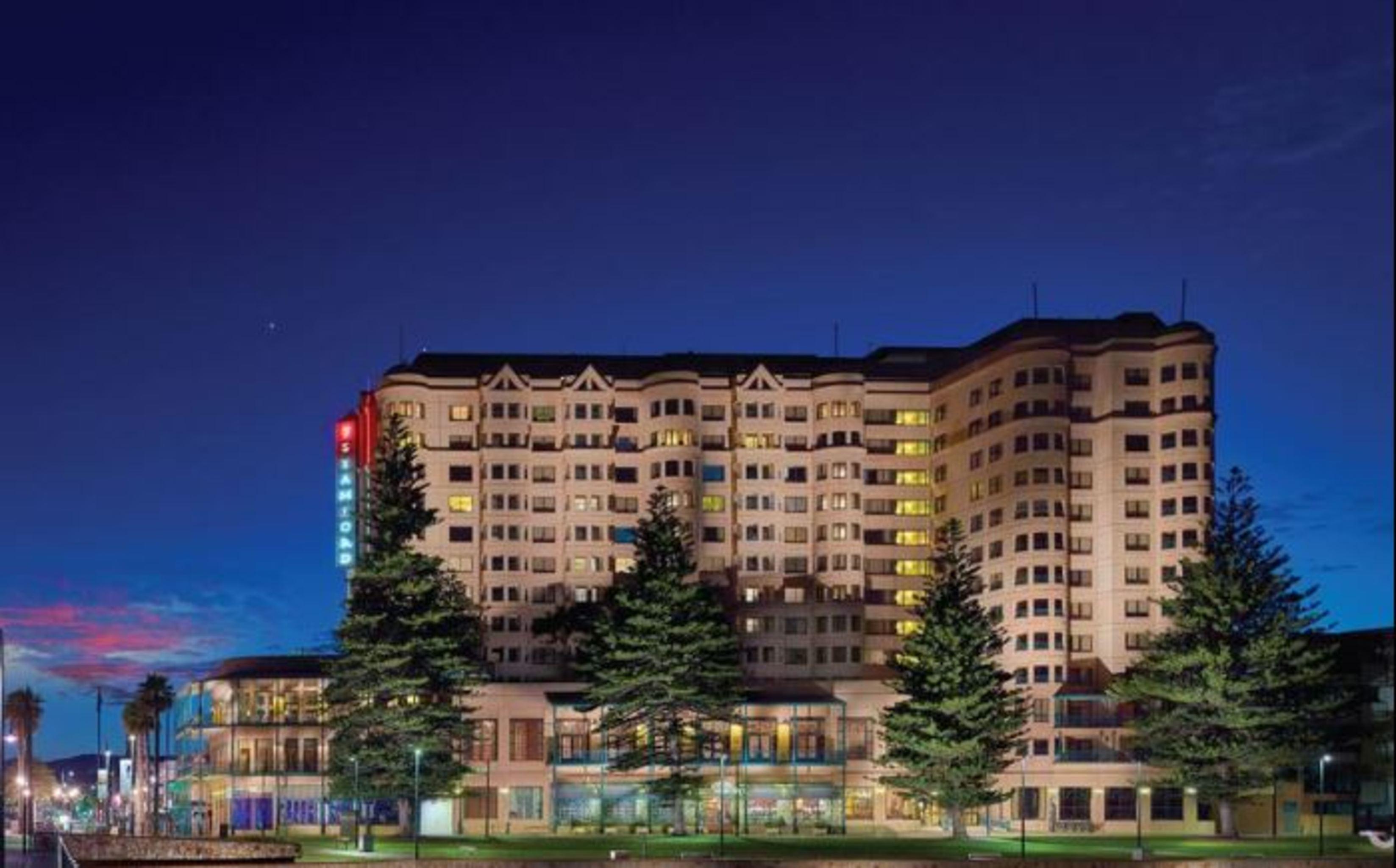 Stamford Grand Adelaide Hotel Glenelg Bagian luar foto The photo displays a multi-story hotel building at dusk, illuminated by various lights. The architecture features a mix of modern and traditional design elements, with numerous windows showcasing a warm glow. Surrounding the hotel are several tall tr