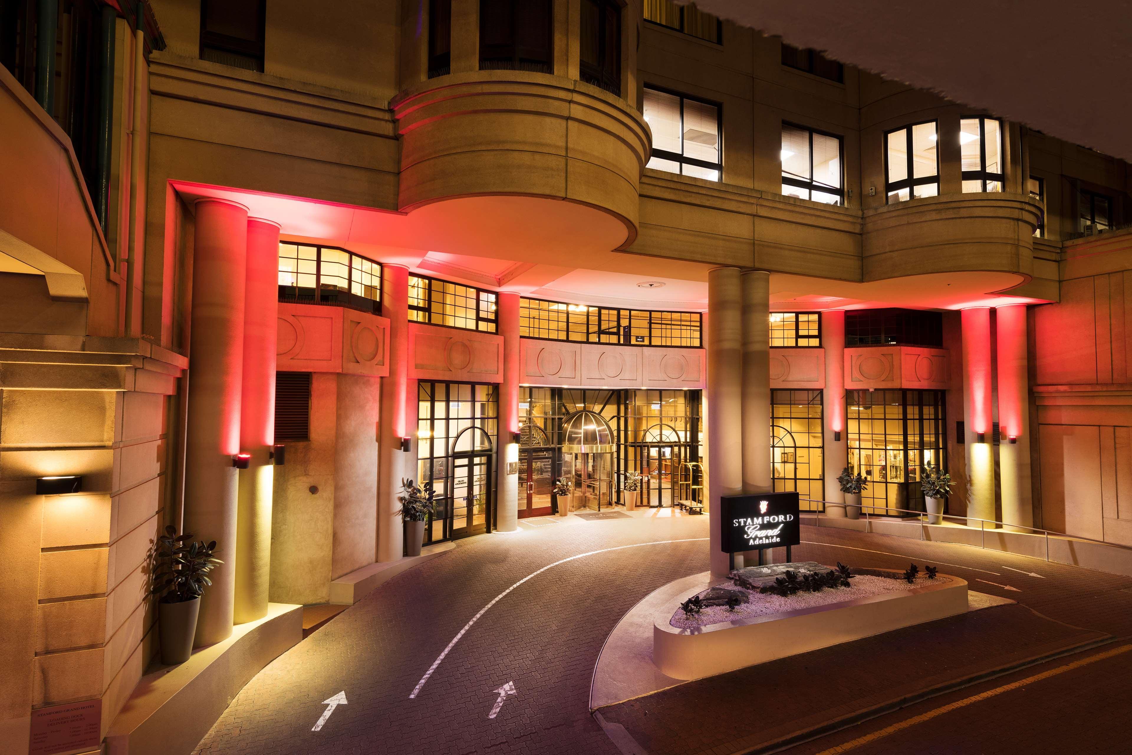 Stamford Grand Adelaide Hotel Glenelg Bagian luar foto The photo shows the entrance of a hotel or luxury building at night. The façade features a modern architectural style with large windows and an elegant entrance. The area is illuminated with red lighting, creating an inviting atmosphere. There appear