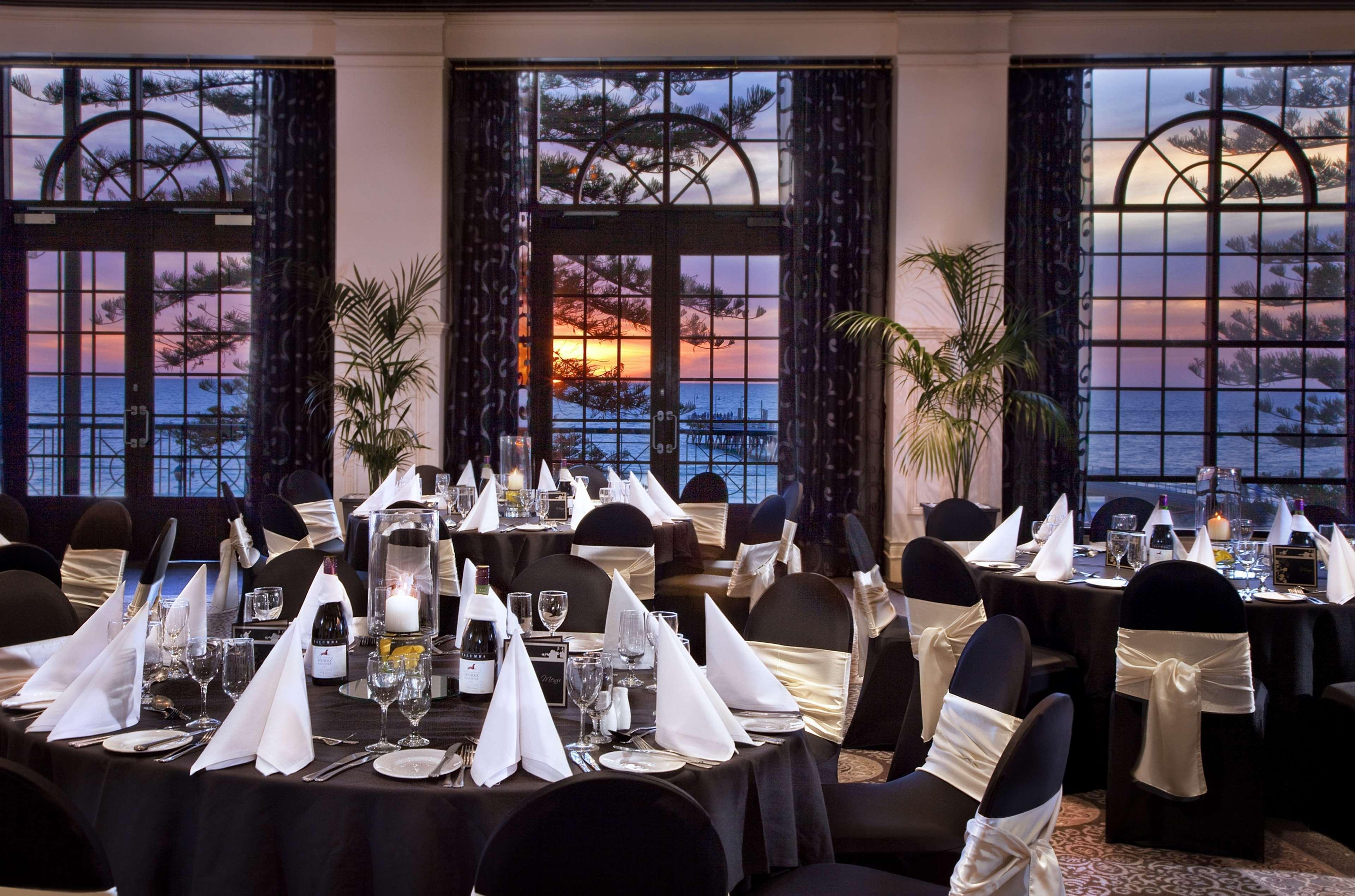 Stamford Grand Adelaide Hotel Glenelg Restoran foto The photo shows an elegantly decorated dining room with a beautiful view. The tables are set with black tablecloths and white napkins folded in a triangular shape. There are glassware and silverware neatly arranged at each setting. The room is adorne