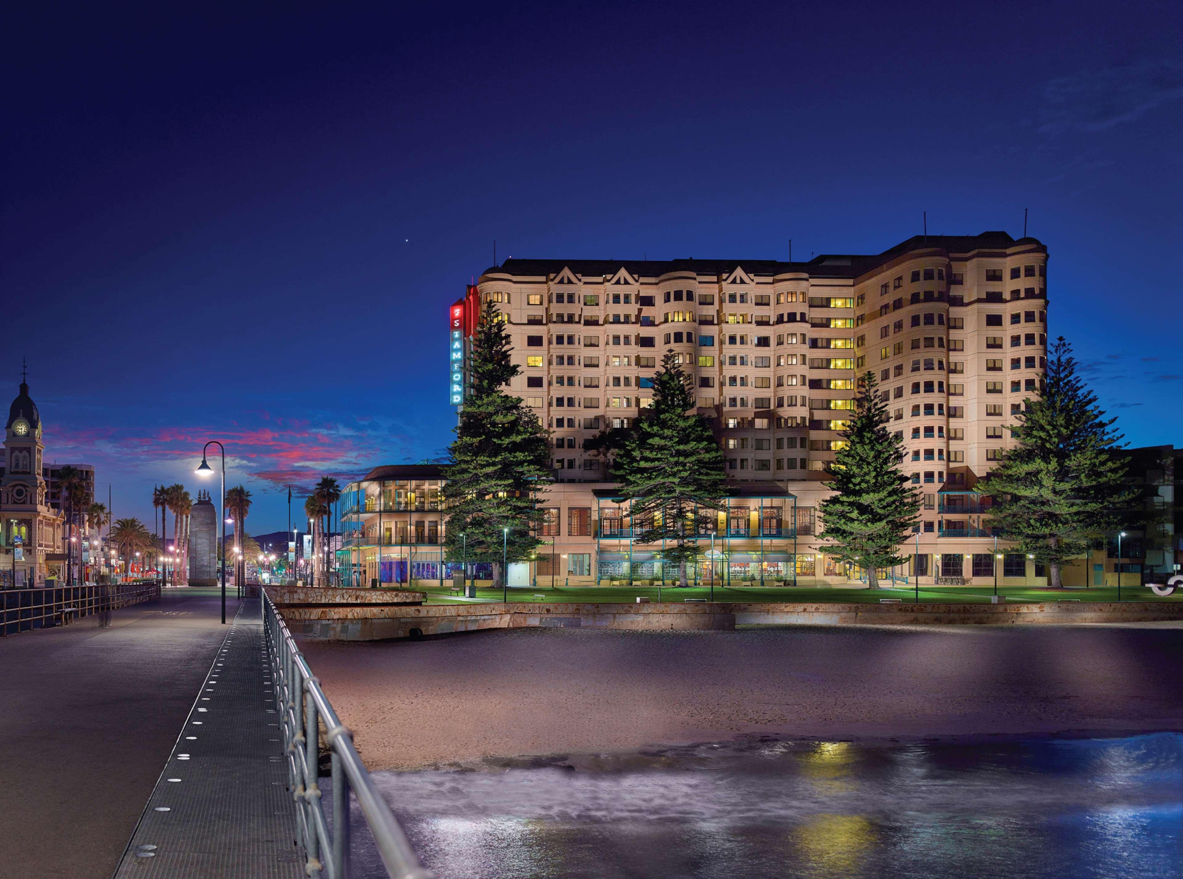 Stamford Grand Adelaide Hotel Glenelg Bagian luar foto The photo shows a waterfront scene at dusk, featuring a large building lit up against a darkening sky. The architecture appears modern and prominent, with several windows illuminated. In the foreground, there is a walkway or pier with fencing along t