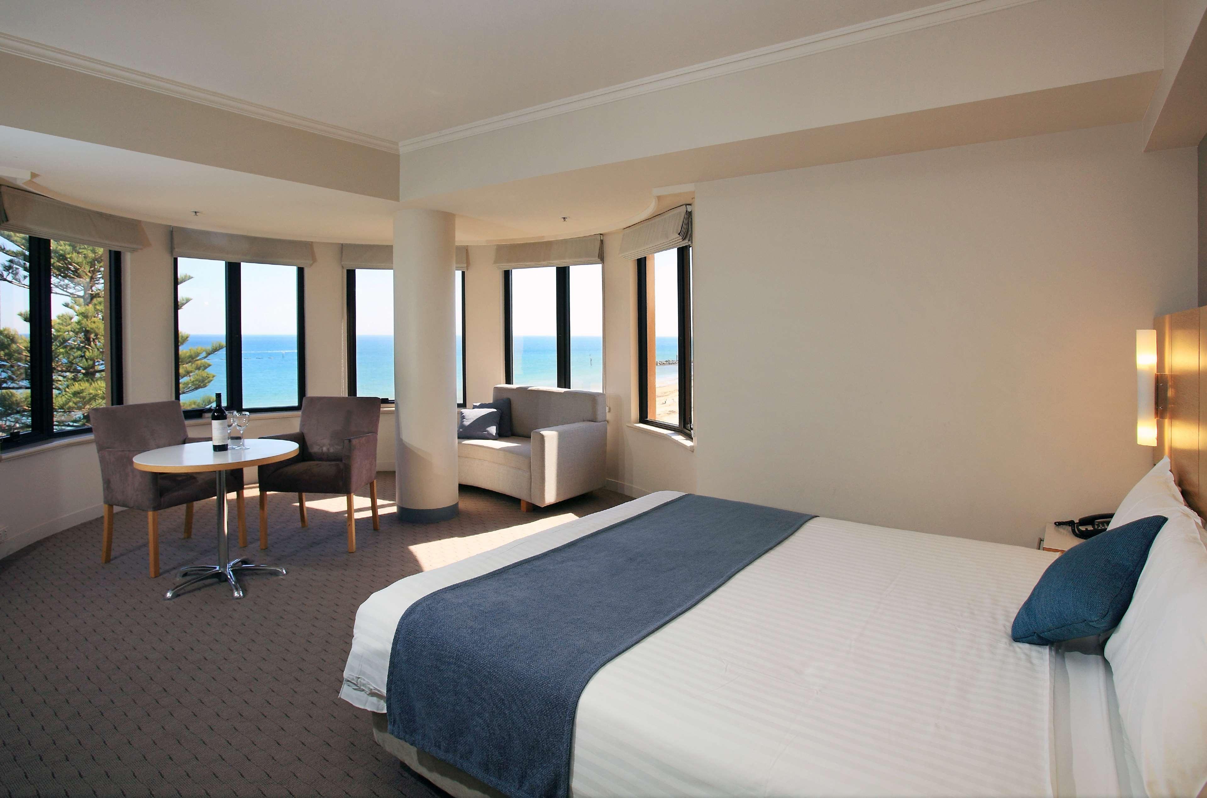 Stamford Grand Adelaide Hotel Glenelg Bagian luar foto The photo shows a well-lit hotel room featuring large windows that offer a view of the ocean. The room is arranged with a comfortable bed covered in white linens and a navy blue throw blanket. There is a small table and chairs set up beside the windo