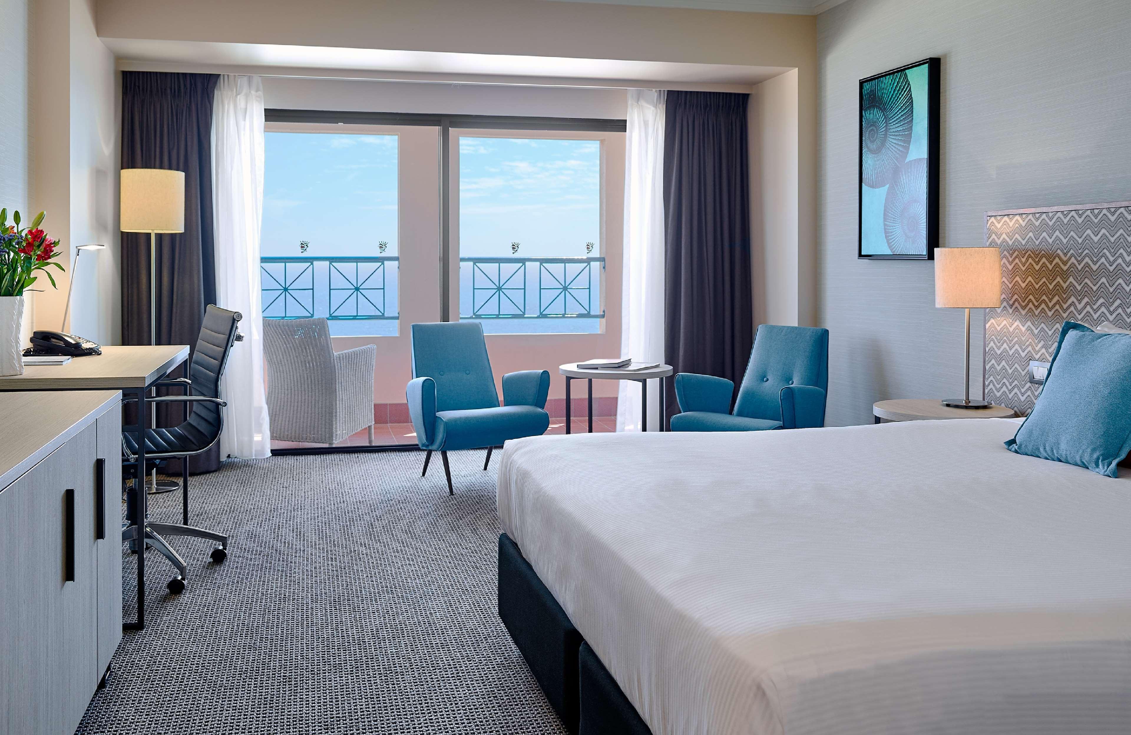Stamford Grand Adelaide Hotel Glenelg Bagian luar foto The image shows a hotel room featuring a large bed with a white duvet. There are two bright blue chairs and a small table near a large window that provides a view outside, possibly to a balcony. The room includes a workspace with a desk and an office