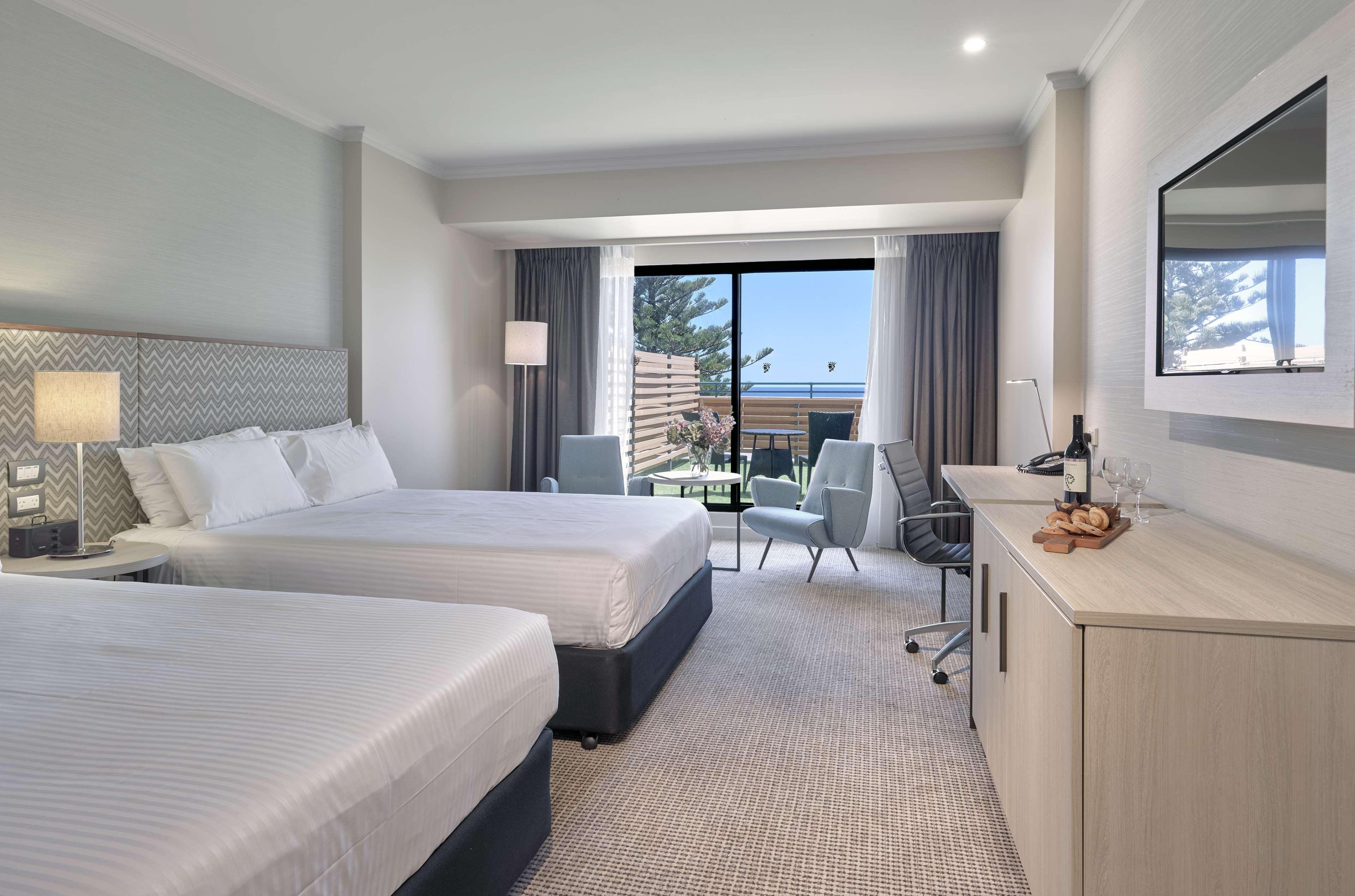 Stamford Grand Adelaide Hotel Glenelg Bagian luar foto The photo shows a modern hotel room featuring two beds with white linens. There's a large window that provides natural light and a view of a balcony area. The room includes two chairs and a small table, creating a cozy sitting area. A television is m