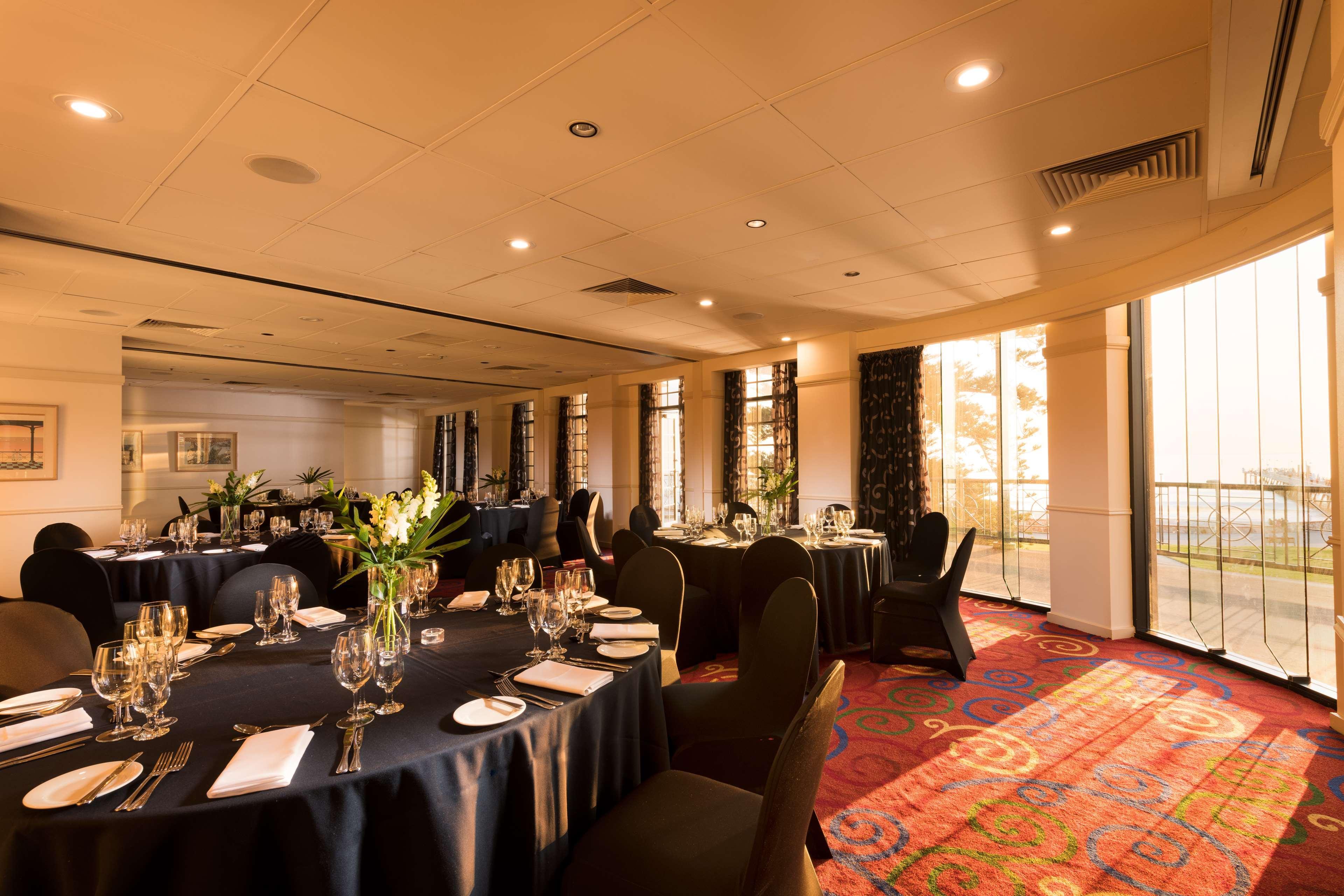 Stamford Grand Adelaide Hotel Glenelg Bagian luar foto The photo shows a well-decorated event space or dining area. There are several round tables draped in dark tablecloths, each set with elegant dinnerware, including glasses and cutlery. The room features large windows that allow natural light to flood