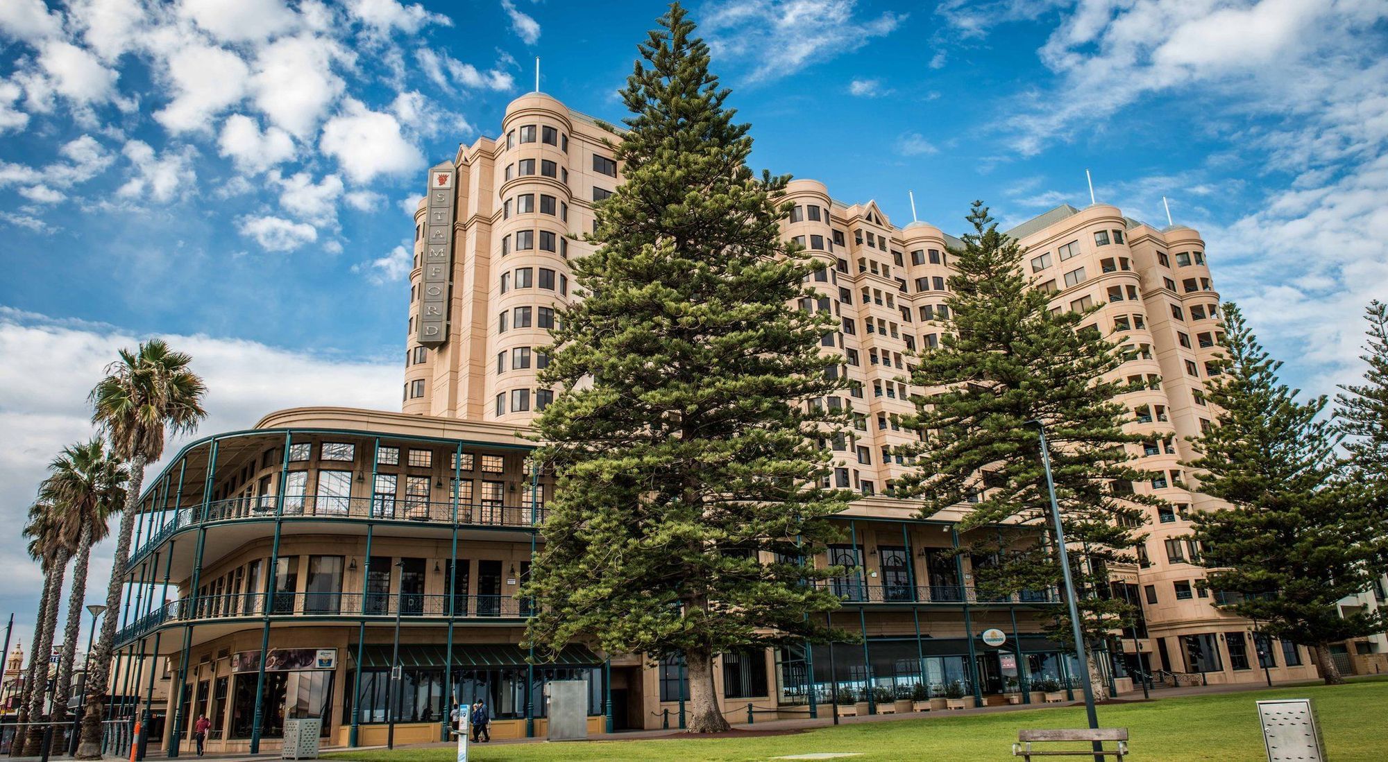 Stamford Grand Adelaide Hotel Glenelg Bagian luar foto The image shows a large building with a distinctive architectural style, characterized by a tan or beige color. The structure appears to have multiple floors and rounded edges. In front of the building, there are several tall green trees, which add t