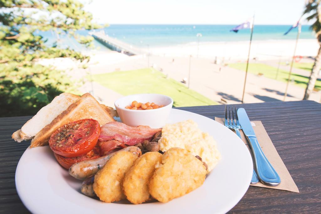 Stamford Grand Adelaide Hotel Glenelg Bagian luar foto The photo shows a plate of breakfast food set against a beachside backdrop. The plate includes several items: crispy bacon, a grilled tomato, sausage links, golden hash browns, and a small serving of baked beans. There is also a piece of toast on the