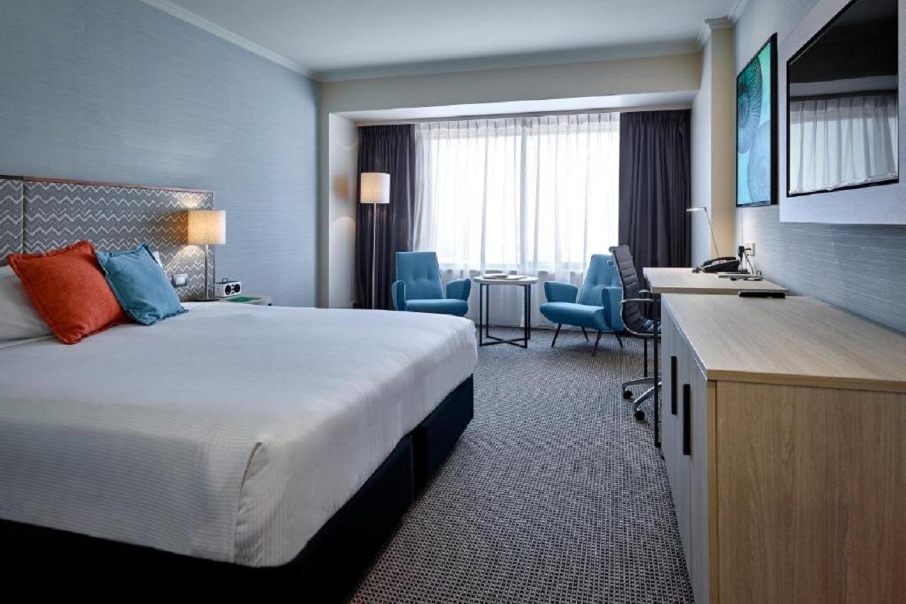 Stamford Grand Adelaide Hotel Glenelg Bagian luar foto The photo shows a modern hotel room with a clean and minimalist design. In the center, there is a large, neatly made bed with white linens and two decorative pillows, one orange and one blue. To the right, there is a small wooden cabinet with a telev